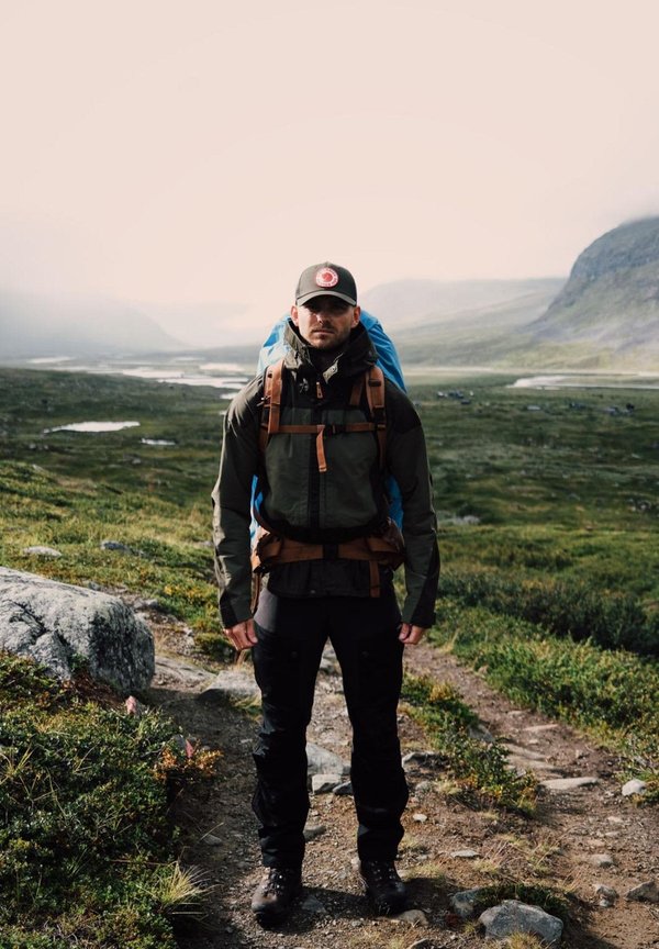 Grön och svart jacka med bruna remmar, svarta byxor och en blå ryggsäck. Personen bär en svart keps med en röd logotyp. Stenlagd väg och grönska.