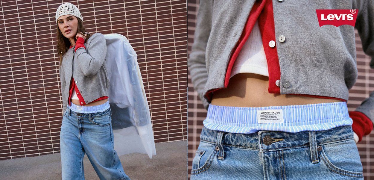 Donna con abiti casual a strati, cappello bianco di maglia e borsa, in piedi davanti a una parete di piastrelle marroni.