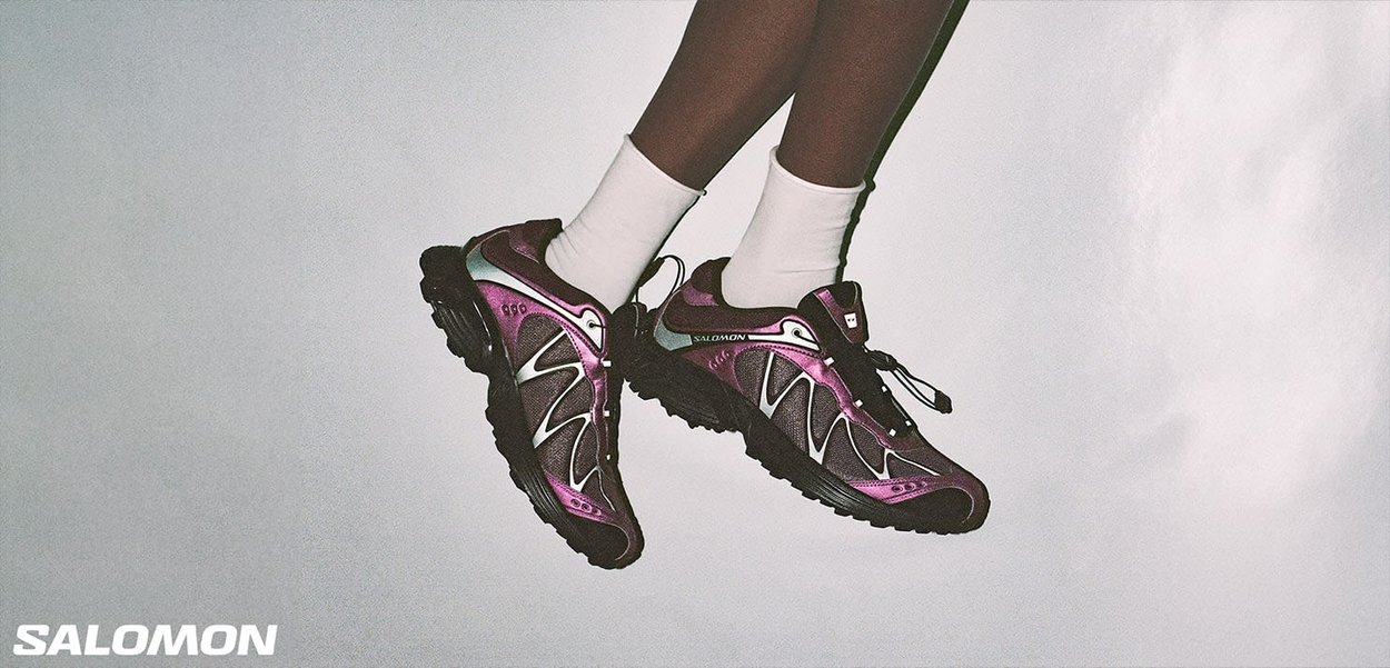 Zapatillas deportivas moradas y negras con materiales de malla y sintéticos, que cuentan con una suela gruesa, acentos reflectantes y un diseño de cordones.