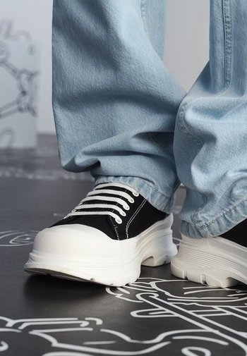 Schwarze Leinenschuhe mit weißen Gummisohlen und Schnürsenkeln, getragen mit hellblauen, locker sitzenden Jeanshosen. Flache Oberfläche unter den Schuhen.
