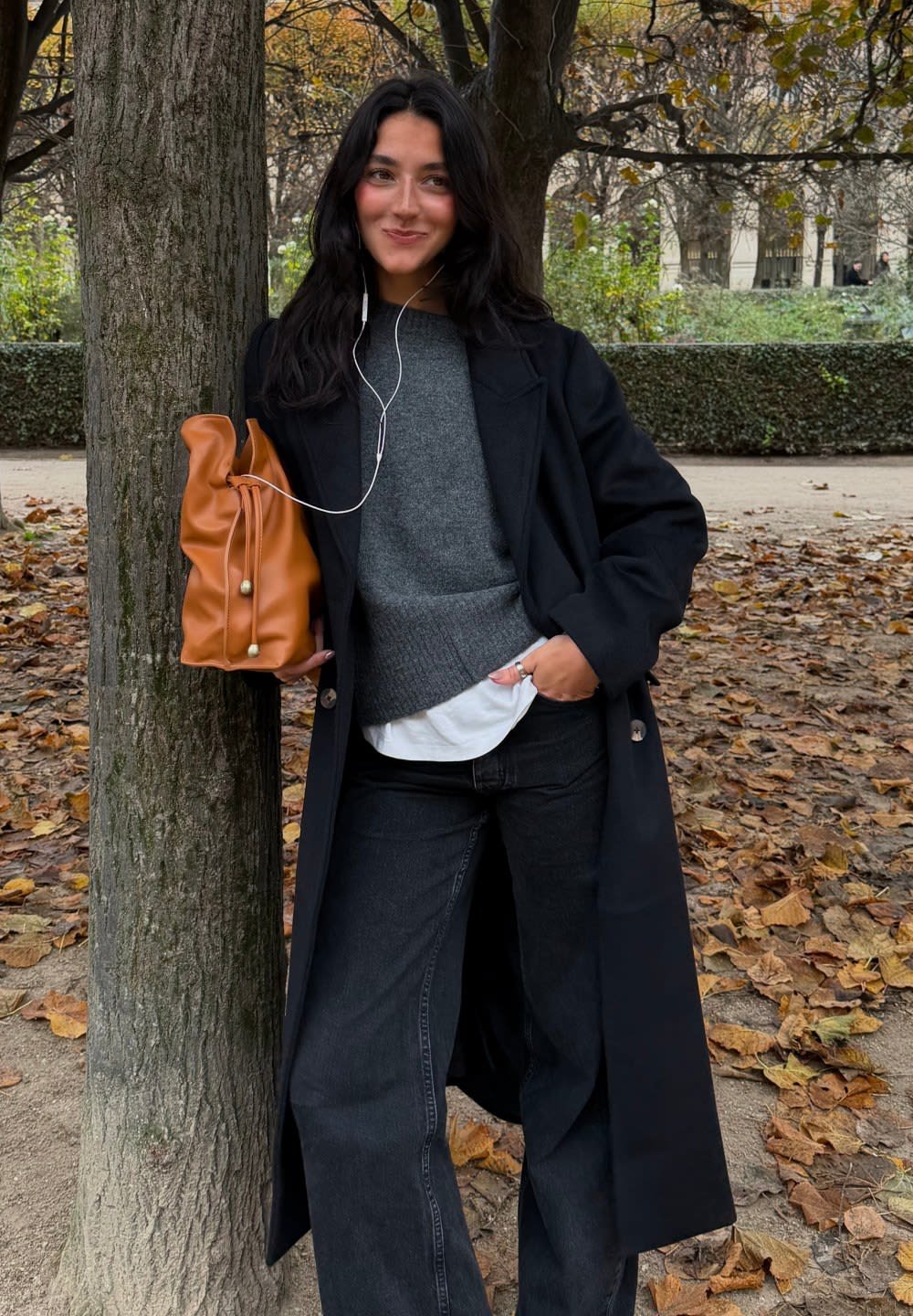 Abrigo largo negro, suéter gris, camisa blanca, vaqueros oscuros de pierna ancha, bolso marrón texturizado con cordón, de pie junto a un árbol con hojas de otoño.
