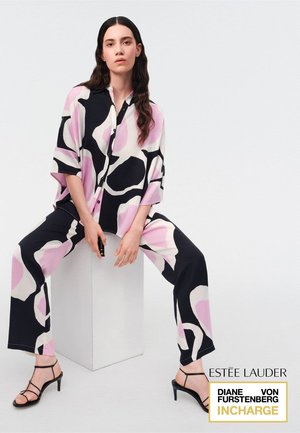 Donna con capelli lunghi e scuri seduta su un blocco bianco, indossa una camicia larga con motivo astratto nero, bianco e rosa e pantaloni coordinati, con tacchi neri.