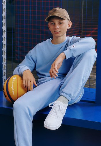 Hellblaues Sweatshirt und Jogginghose aus weichem Stoff, kombiniert mit weißen Turnschuhen. Hält einen orangefarbenen Basketball mit schwarzen und gelben Akzenten.