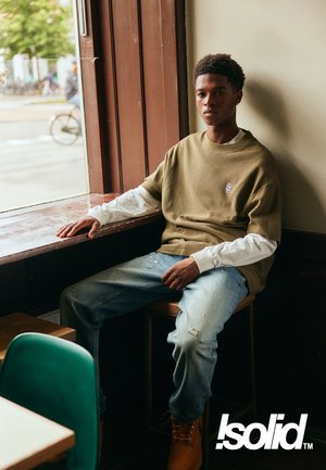 Olijf groene ribbeltrui over een witte lange mouw, versleten lichtblauwe spijkerbroek en bruine laarzen; zittend bij een raam in een café.