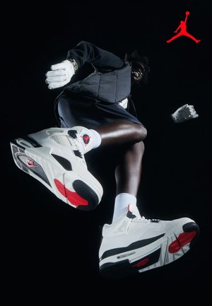 Osoba nosząca białe i czerwone sneakersy Air Jordan, czarne spodenki, czarną kamizelkę, białe rękawiczki i okulary przeciwsłoneczne, skacząca na ciemnym tle.