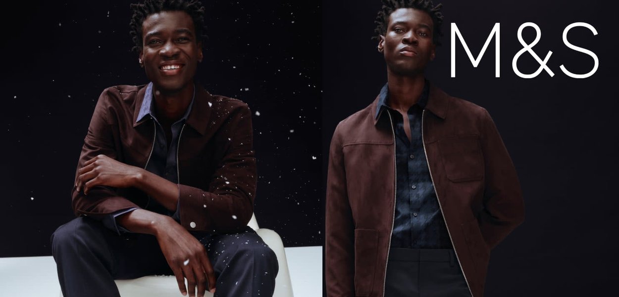 Brązowa zamszowa kurtka na ciemnej koszuli na guziki, w połączeniu z ciemnymi spodniami. Model ma krótkie, teksturowane włosy na neutralnym tle.