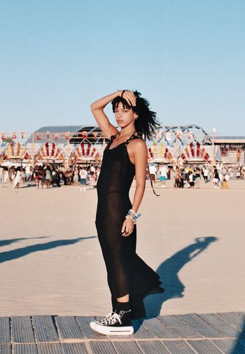 Black sheer maxi dress with thin straps and a fitted silhouette, paired with black high-top sneakers and blue bracelets against a beach background.