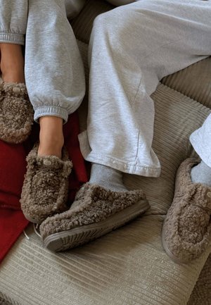 Braune, flauschige Hausschuhe mit einer strukturierten Außenseite, Gummisohlen und einem Etikett. Kombiniert mit grauen Jogginghosen und einer roten Decke.