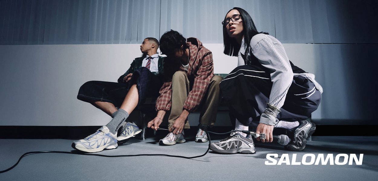 Trois jeunes portant des baskets Salomon, l'un accroupi avec un micro, un autre attachant ses lacets, et le troisième assis contre un mur.