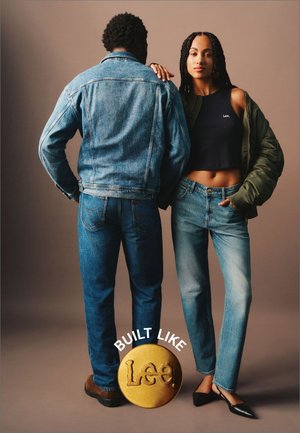 Jeansjakke og jeans i forskellige nuancer af blå, parret med en sort cropped top og en olivengrøn bomberjakke. Stort guld rundt logo med tekst nedenfor.