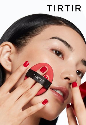 Red silicone makeup applicator with a black strap, held by a hand with red nail polish on a model's face, showcasing smooth skin texture.