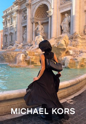 Vestido negro sin mangas con una falda de capas, combinado con un bolso negro con herrajes dorados, contra el fondo de una fuente de piedra.
