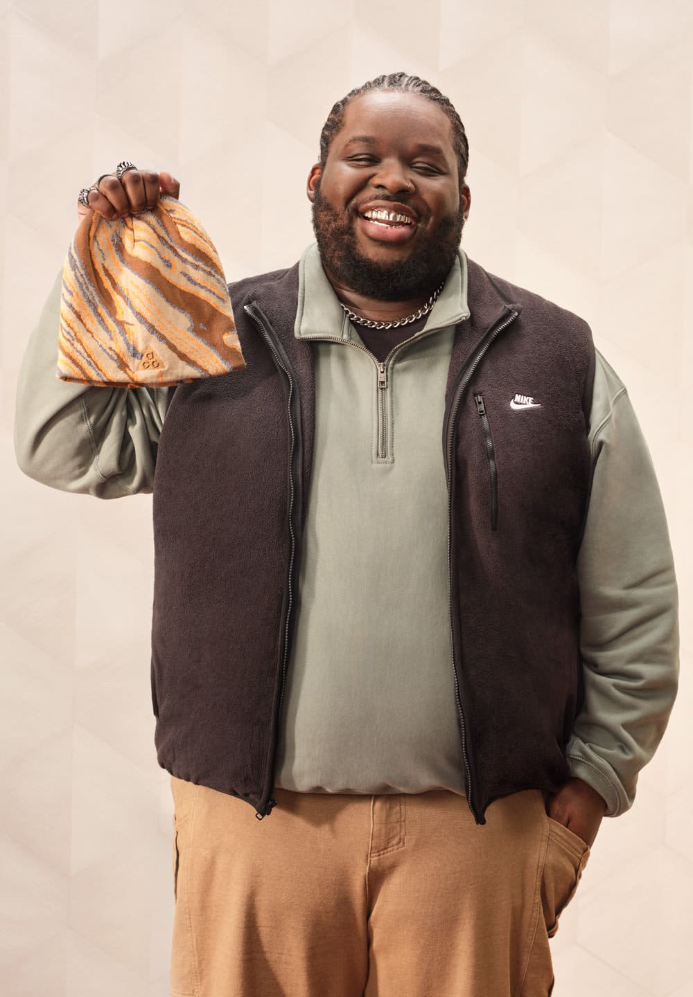 Gilet in pile nero sopra una maglia verde a maniche lunghe, che tiene un berretto marmorizzato arancione e blu. Sullo sfondo è presente un motivo geometrico chiaro.