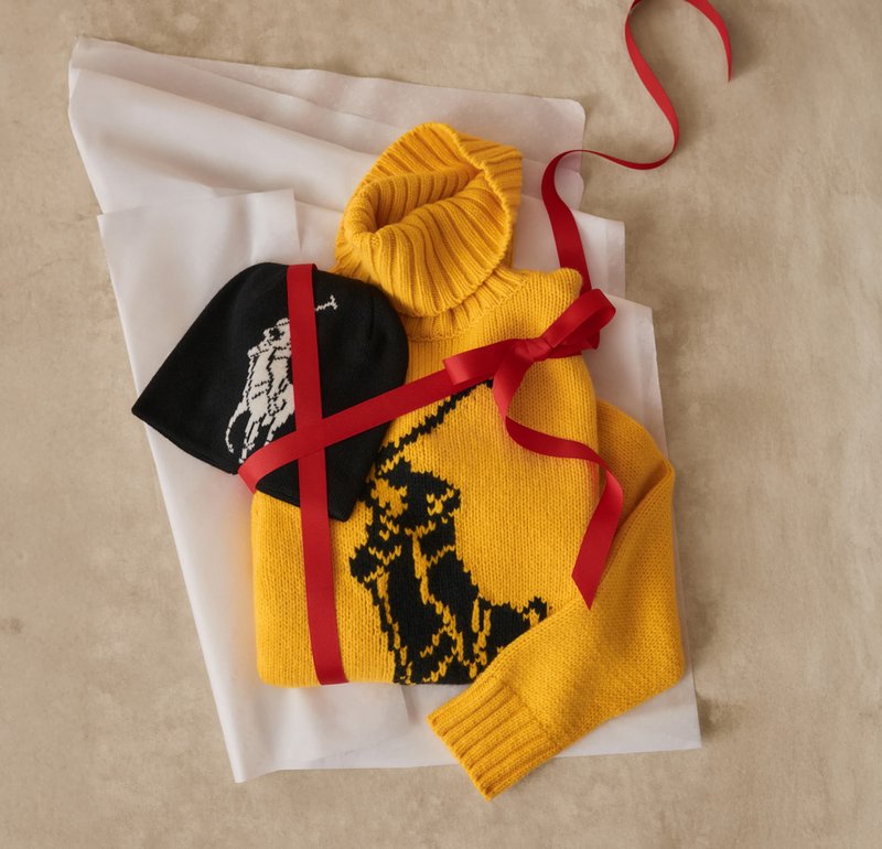 Gul strikket turtleneck-genser med svart dyremønster, kombinert med en svart lue med et hvitt grafisk motiv, bundet med et rødt bånd.