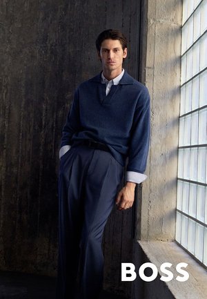 Jonge man in een marineblauwe trui en broek leunt tegen een betonnen muur naast een groot glazen raam in een schemerige ruimte met het BOSS-logo.