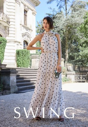 Frau in einem ärmellosen weissen Maxikleid mit schwarzen Punkten steht draussen neben Steinstufen und hält eine metallische Clutch.