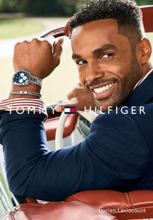 Lächelnder Mann im marineblauen Blazer sitzt auf einem roten Ledersitz im Auto, trägt silberne Uhr und Armband, schaut über die Schulter zurück und hält das Lenkrad.