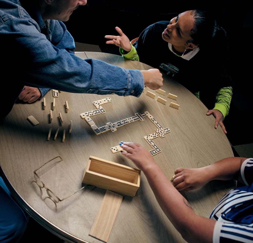 Mediniai domino yra sutvarkyti ant apvalaus medinio stalo. Šalia yra šviesios spalvos medinė dėžė, o stalo krašte stovi stiklinės.