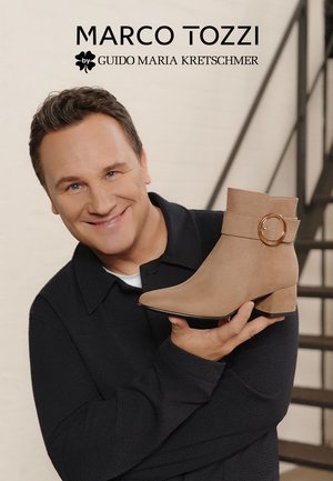 Beige suede ankle boot with a pointed toe, flat heel, and a wide strap featuring a circular metal accent. Displayed against a neutral background.