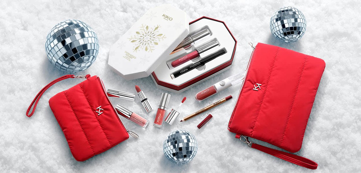 Czerwony pikowany kosmetyczny woreczek ze srebrnymi okuciami, otoczony kosmetykami oraz dekoracyjnym białym pudełkiem z logo KIKO. Akcent w postaci kuli dyskotekowej.