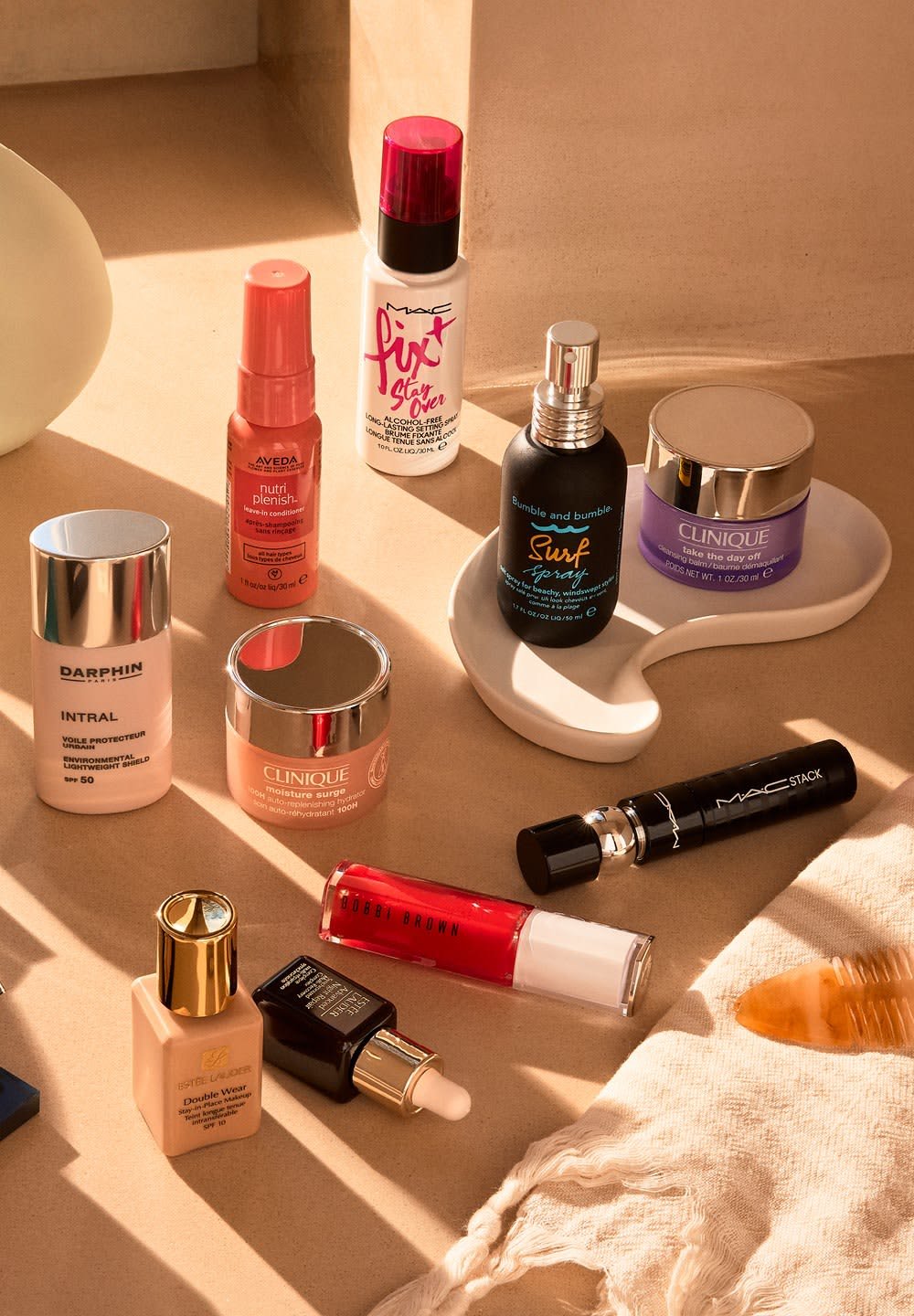 Dieci prodotti cosmetici disposti su una superficie neutra. Caratteristiche includono varie forme, colori e finiture: creme, spray e sieri in packaging distintivi.
