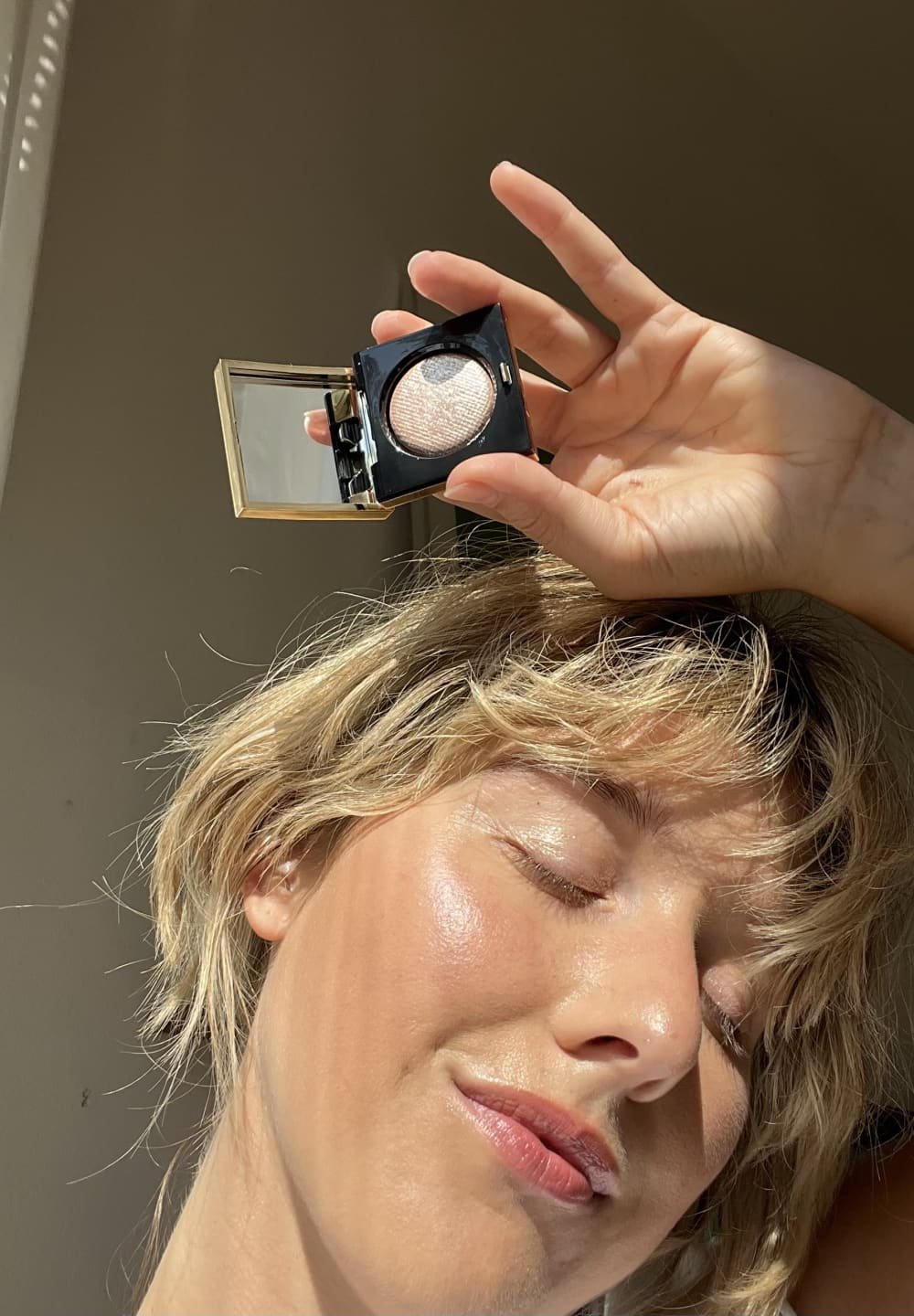 Femme les yeux fermés tenant un petit miroir de maquillage ouvert avec un illuminateur scintillant près de sa tempe, sous une douce lumière naturelle.