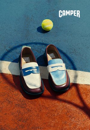 Mocasines de cuero bicolor sobre una cancha deportiva multicolor con una pelota de tenis amarilla con la marca "Camper" cerca.