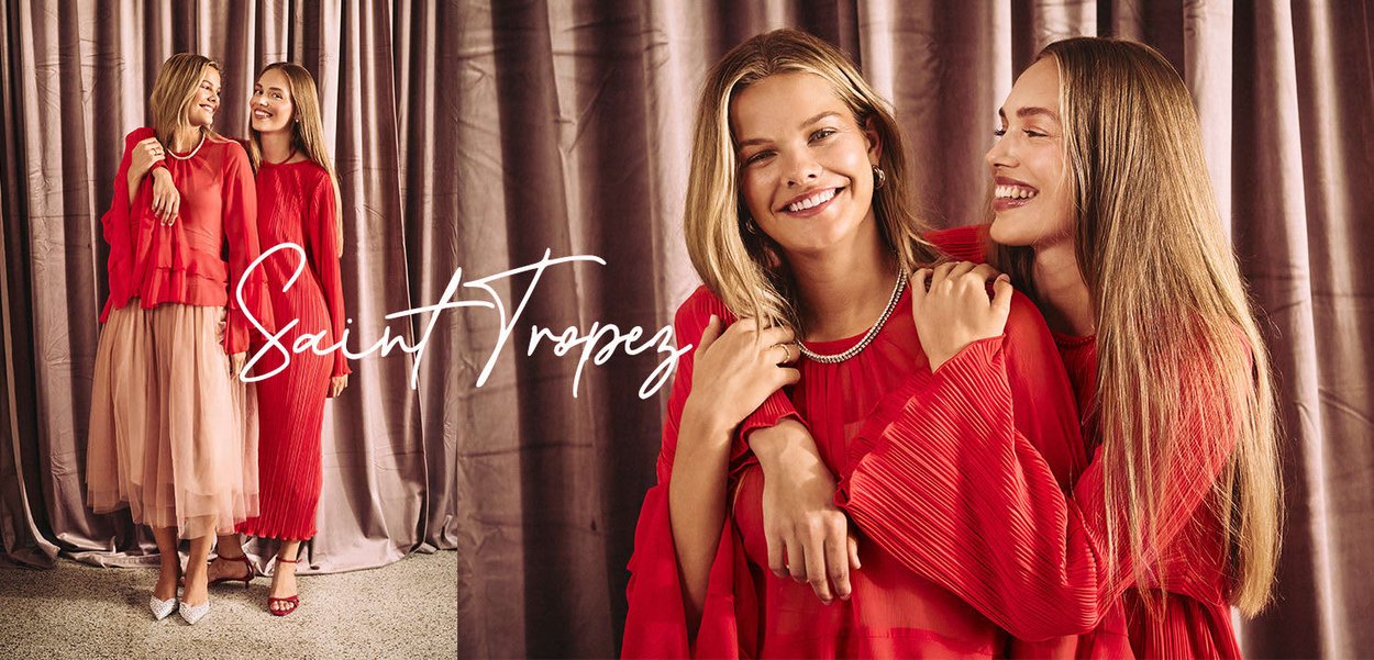 Zwei Frauen tragen lebhafte rote Outfits; eine in einem plissierten Kleid, die andere in einer gerüschten Bluse mit einem Tüllrock. Weiche Stoffe, Lächeln und ein neutraler Hintergrund.