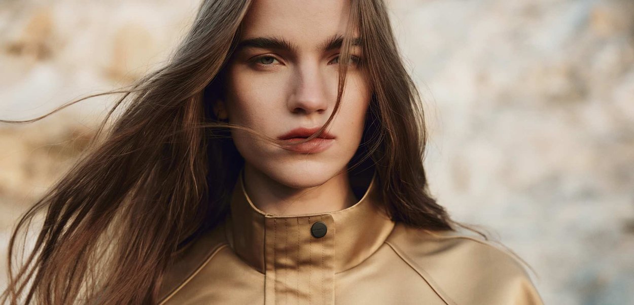Młoda kobieta o długich brązowych włosach, ubrana w beżową kurtkę, patrząca prosto w obiektyw na rozmytym, zewnętrznym tle.