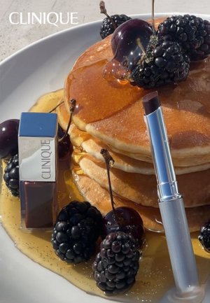 Stack of golden pancakes drizzled with syrup, surrounded by blackberries and cherries, with a silver Clinique lipstick tube placed nearby.
