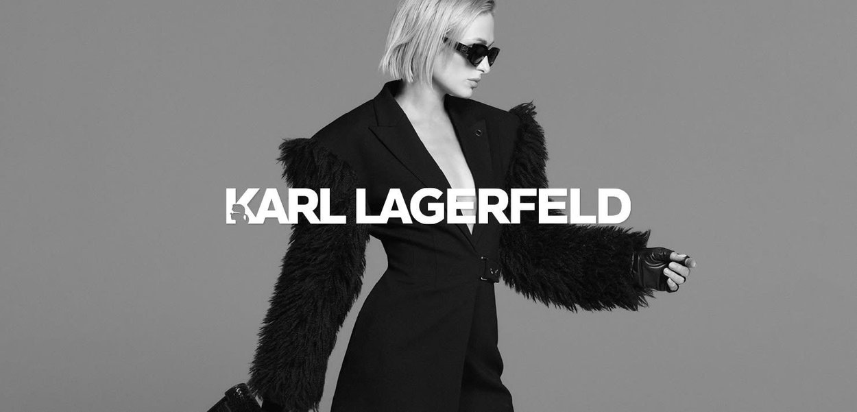 Black tailored jacket with fur sleeves, featuring a fitted silhouette. Model wearing oversized sunglasses and holding a small handbag.
