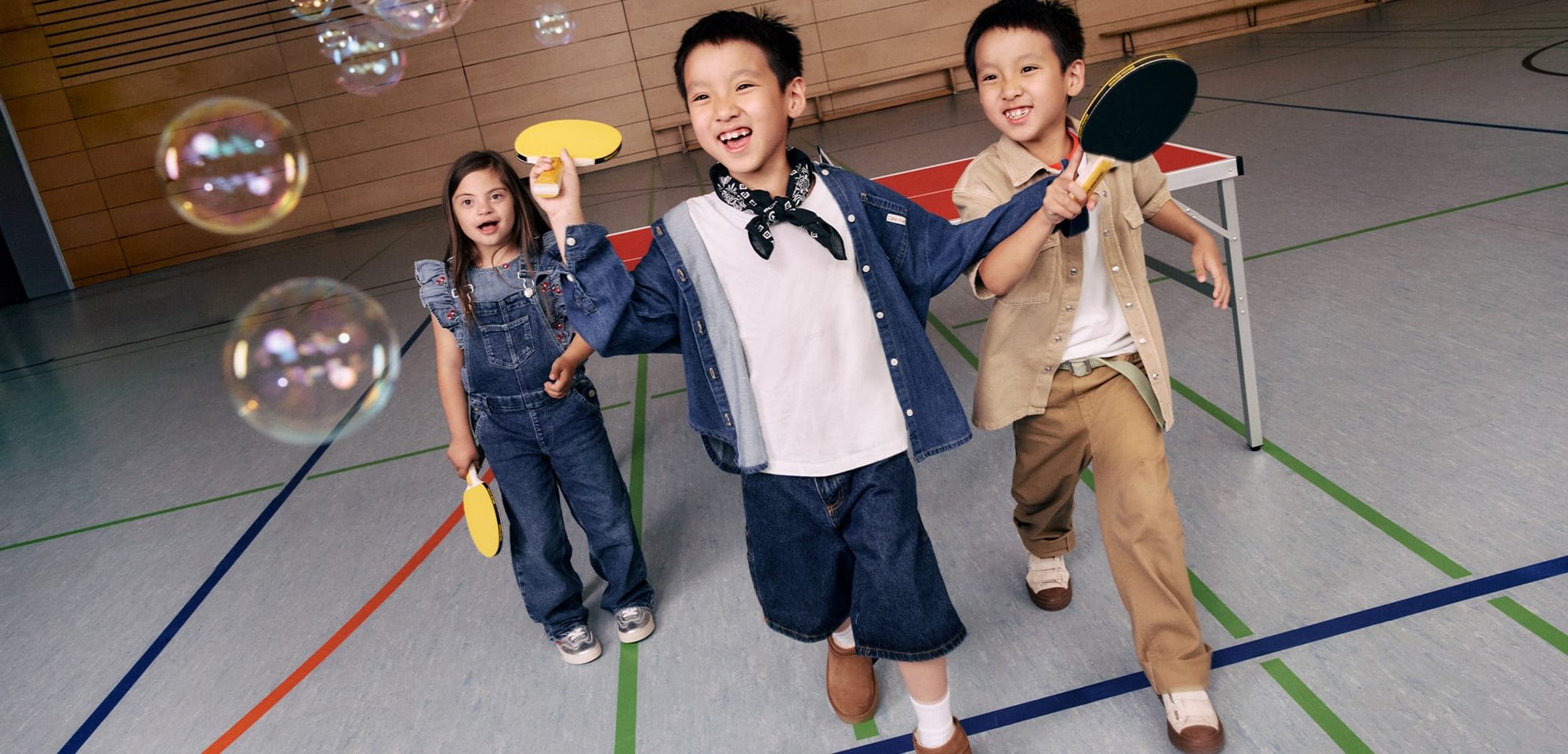 Trois enfants jouent avec des raquettes de ping-pong jaunes dans une salle de sport. Salopettes en denim, chemises claires et pantalons beiges. Des bulles flottent dans l'air.