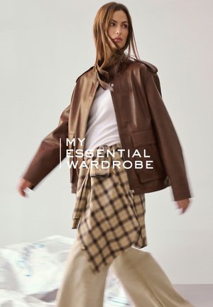 Frau trägt braune Lederjacke, weißes Oberteil, beige karierter Rock, geht mit wehenden Haaren, Text lautet „Meine unverzichtbare Garderobe.“