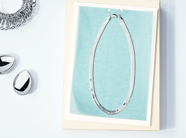 Ασημένιο κολιέ με λεία, επίπεδη σχεδίαση, τοποθετημένο σε φόντο σε απόχρωση aqua. Δύο λαμπερά ασημένια σκουλαρίκια κοντά, σε σχήμα δάκρυ.