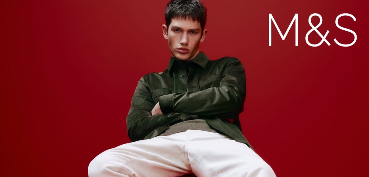 Zielonkawo-brązowa kurtka z kołnierzykiem na jasnej koszuli, w połączeniu z białymi spodniami, pozująca na tle głębokiego czerwonego tła. Logo marki „M&S” po prawej stronie.