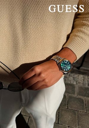 RVS-horloge met een turquoise wijzerplaat, zilveren en gouden armband. Persoon houdt zwarte zonnebril vast in een gestructureerde beige sweater.