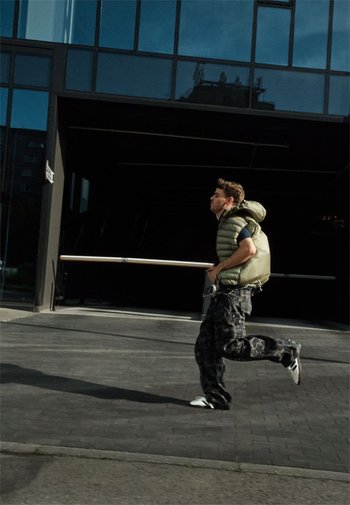 Olijfgroene puffer vest, oversized ontwerp, met een capuchon. Gepaard met donkere camouflagebroek en witte sneakers, rennend op een pavement.