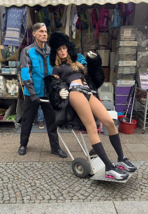 Eine Frau in einem schwarzen Pelzmantel und Shorts sitzt auf einem Wagen mit Rädern, während ein Mann in einer blauen Jacke hinter ihr steht. Im Hintergrund sind Taschen und Stoffe zu sehen.