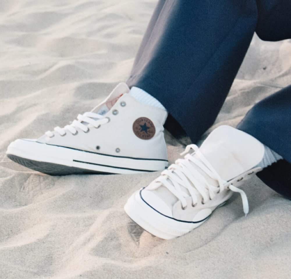 White canvas sneakers feature a round brown logo patch, white laces, black rubber soles, and contrasting toe cap. Worn with navy pants on sand.