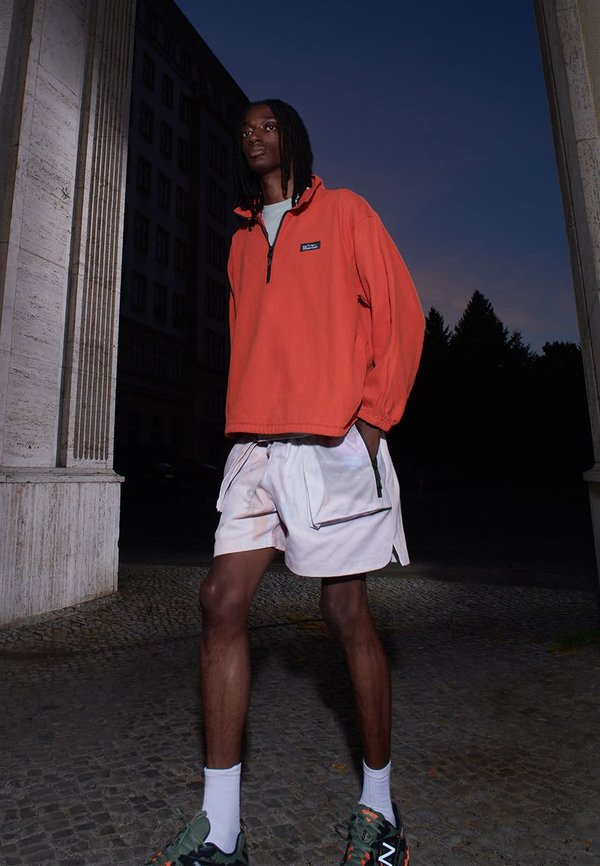 Sudadera de forro polar naranja con media cremallera, combinada con pantalones cortos claros que tienen un bolsillo. El modelo lleva zapatillas verdes y calcetines blancos.