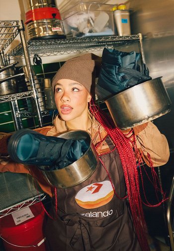 Delantal marrón con logo, sosteniendo ollas metálicas que contienen botas azules. El fondo incluye equipo de cocina en estantes, con azulejos verdes visibles.