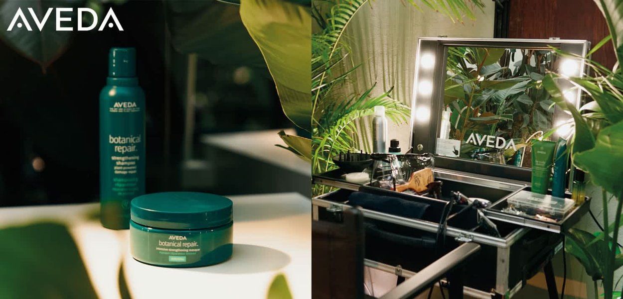 Shampoing et masque réparateurs botaniques Aveda dans un emballage vert, présentés sur un meuble de salle de bains avec miroir éclairé, entourés de feuillage vert et de produits.