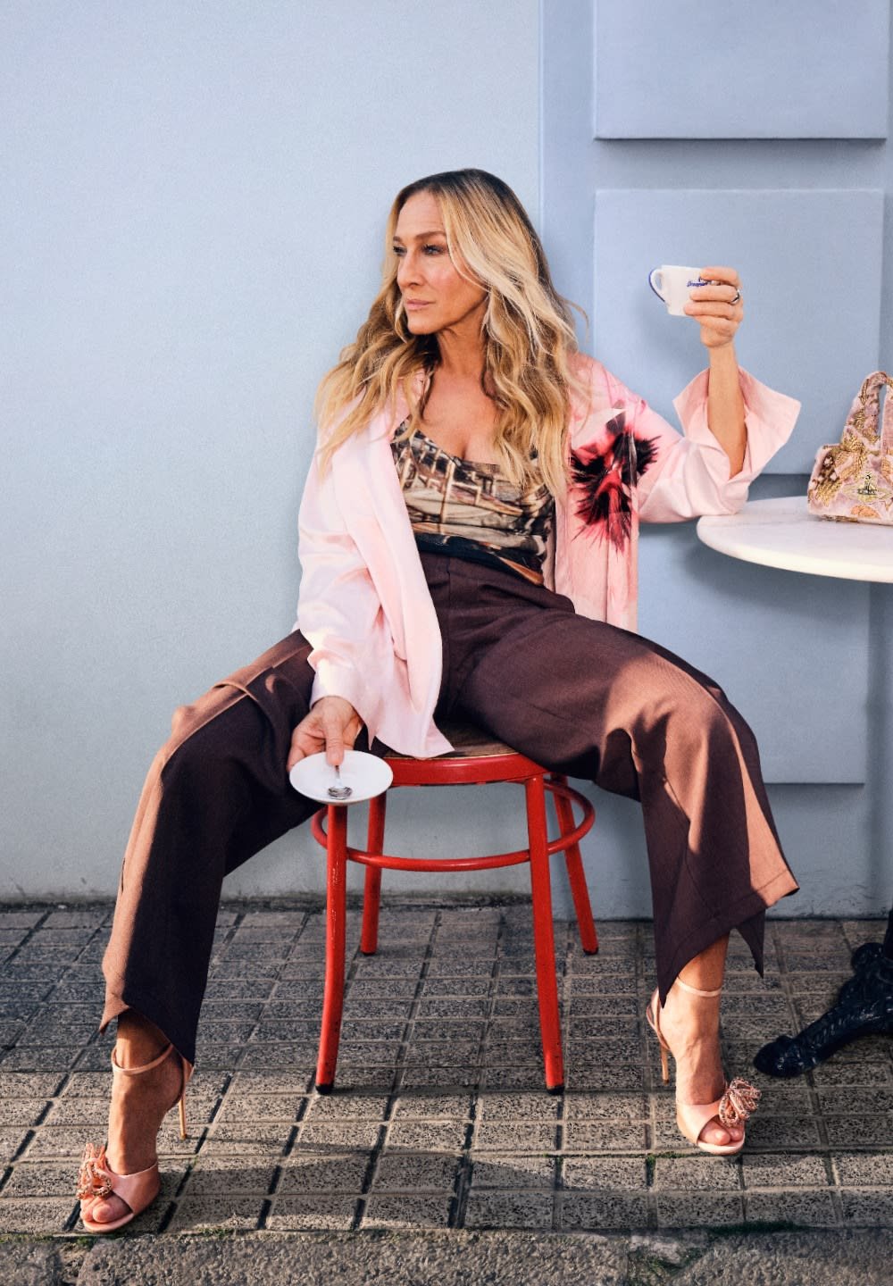 Model u ružičastoj oversized košulji, s uzorkastim topom i smeškim širokim hlačama, drži bijelu šalicu, sjedi na crvenoj stolici.