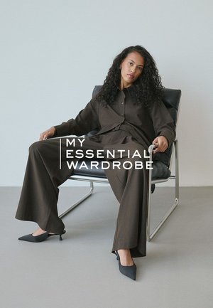 Pantalons larges à côtes brunes avec une chemise à boutons assortie. Des talons hauts complètent la tenue. Chaise noire et fond minimaliste.