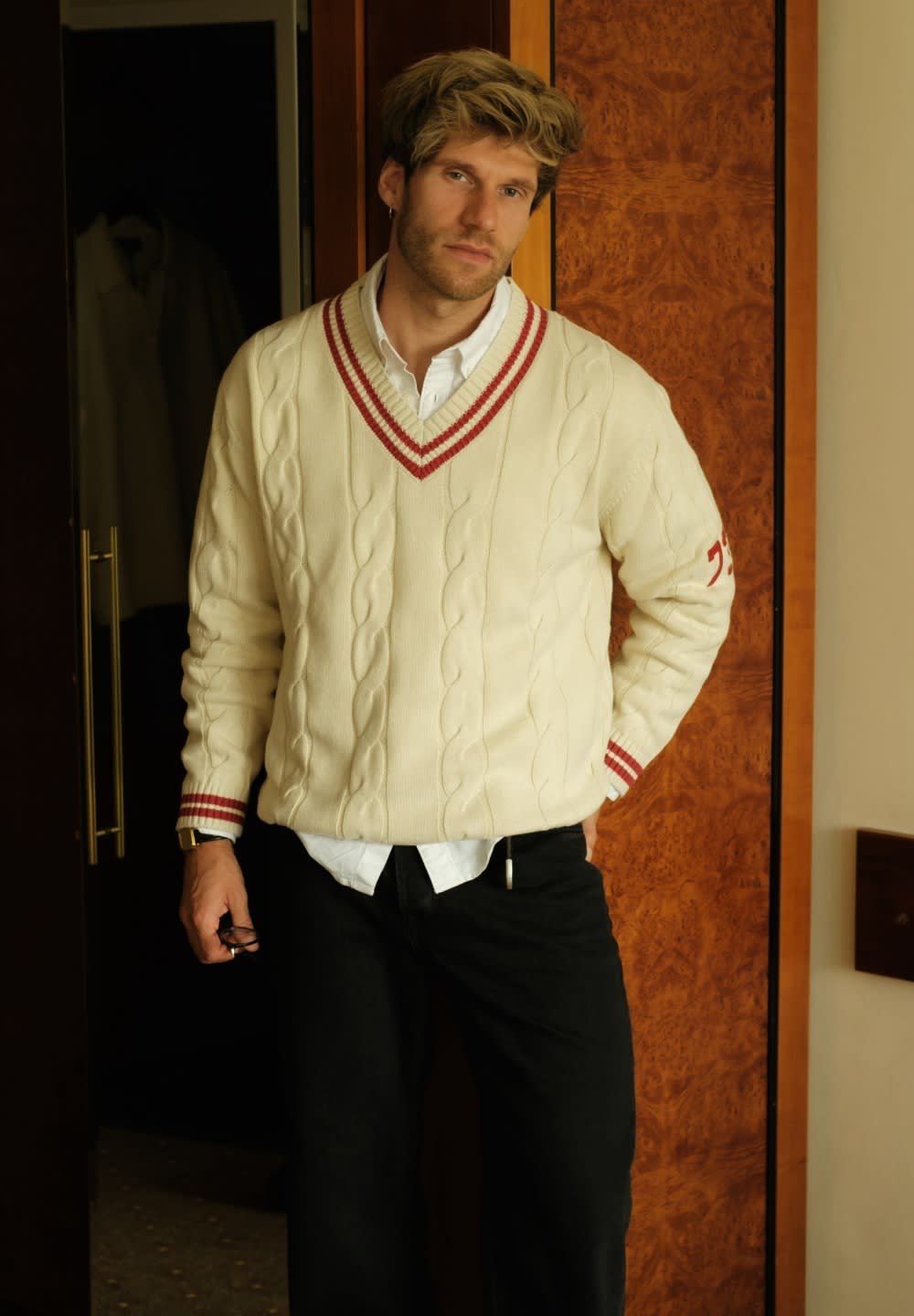 Suéter de punto en crema con ribete en V de rayas rojas, llevado sobre una camisa blanca y pantalones negros. Muestra un patrón de punto texturizado y un ajuste holgado.