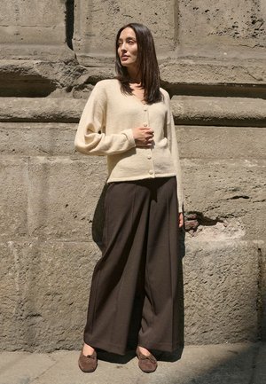 Cream wool cardigan with a v-neck and button closure, paired with wide brown pants and brown suede shoes, set against a stone background.