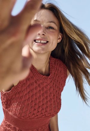 Junge Frau mit langen Haaren lächelt und greift zur Kamera, trägt ein rotes gestricktes ärmelloses Top vor klarem blauem Himmel.