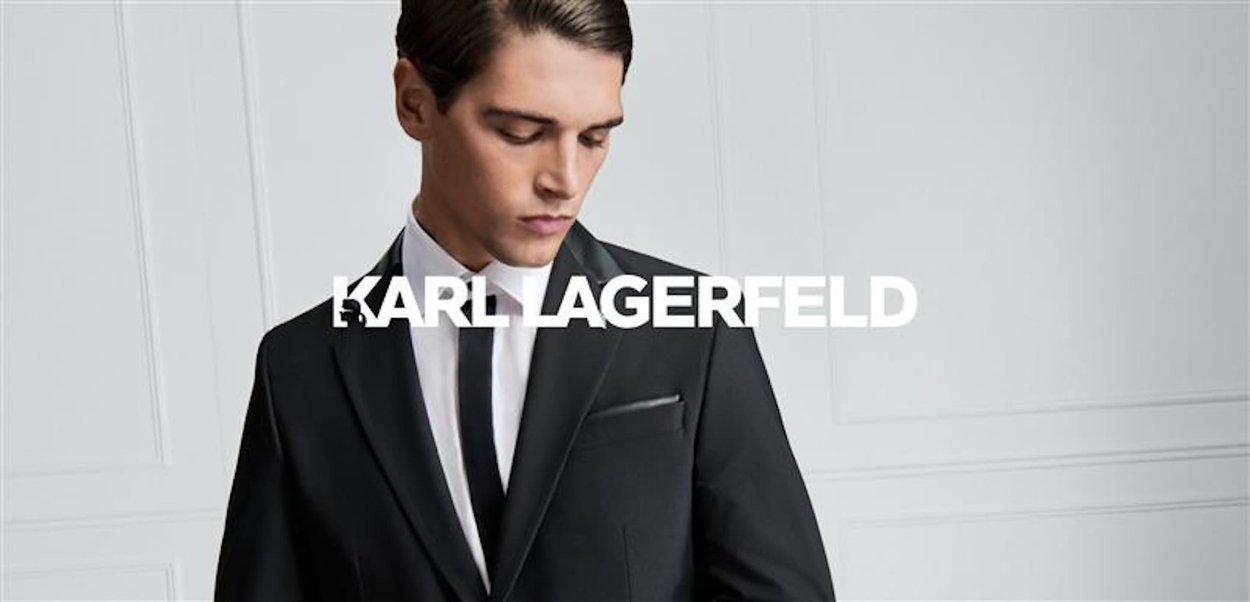 Czarny, szyty na miarę garnitur z satynowym klapem, biała koszula i czarny krawat. Na klapie znajduje się logo "K". Gładka faktura materiału.