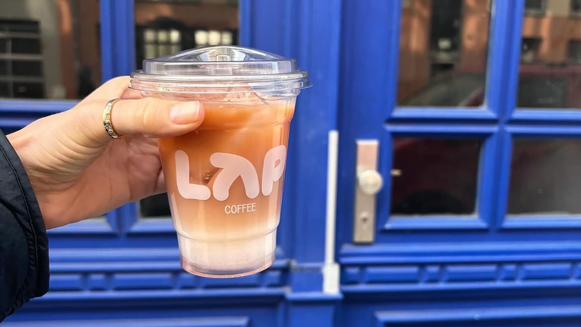 Doorzichtig plastic beker met iced coffee, met een gelaagde bruine en romige kleur. Het etiket toont "LAP COFFEE" in vetgedrukte witte tekst.