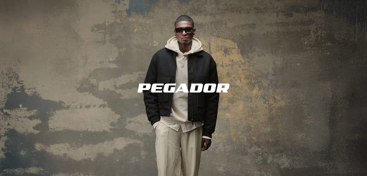 Mann mit schwarzer Jacke, beigem Hoodie und Sonnenbrille steht vor einer strukturierten grauen Wand mit dem "PEGADOR"-Logo in der Mitte.